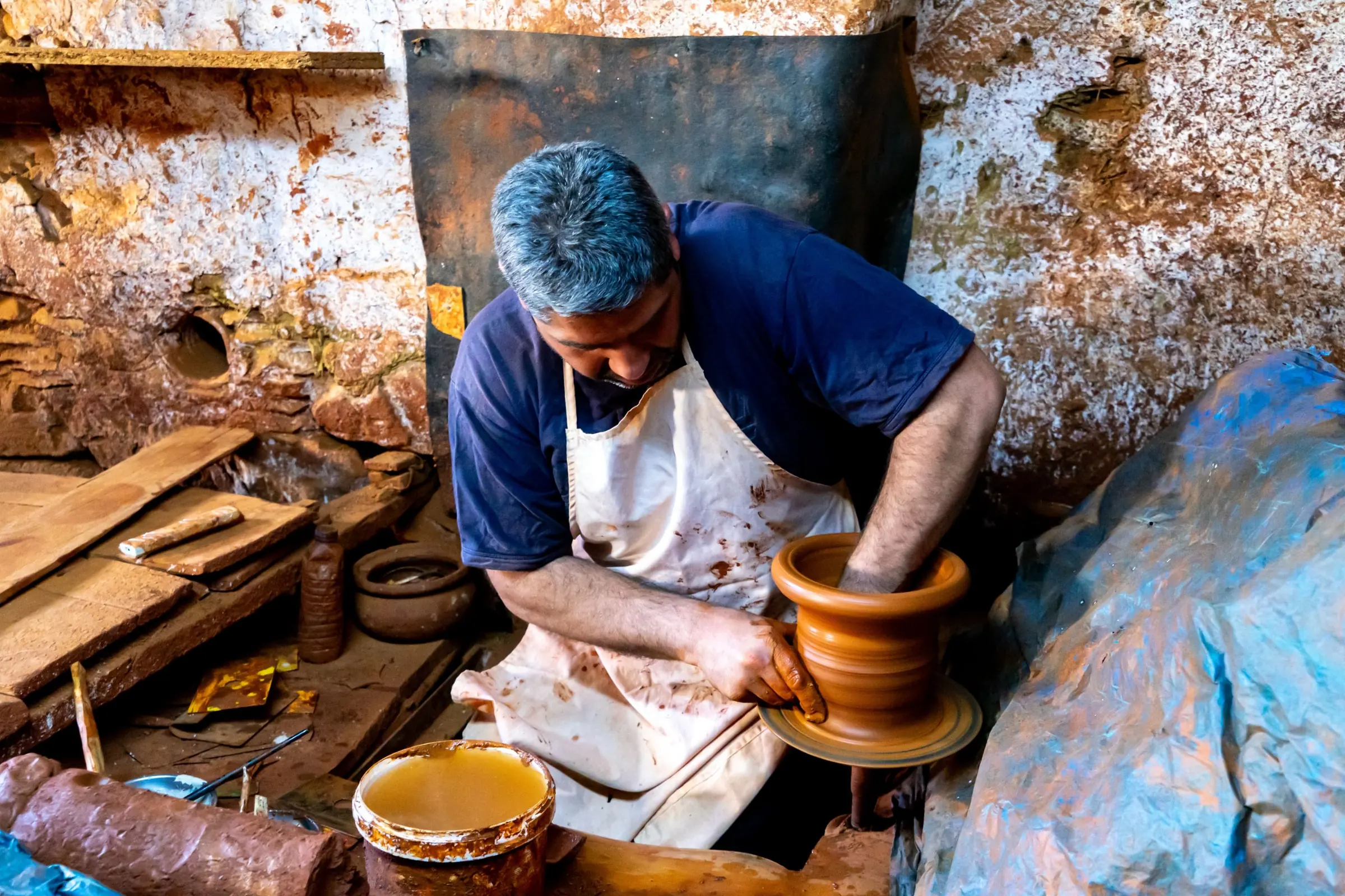Un potier en tablier blanc tourne un vase d'argile sur son tour, dans un atelier aux murs de terre.